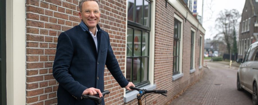 Een afbeelding van een man die buitenstaat met een fiets. Hij draagt een donkerblauwe jas en glimlacht naar de camera.  