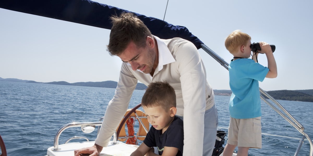 Een man met 2 kinderen op de boot. Een van de kinderen kijkt door een verrekijker richting de zee